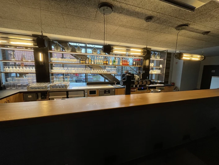 IMG_9237.jpg Moderne Bar mit Holztheke, beleuchteten Glasregalen, Getränkeflaschen und Kaffeemaschine.