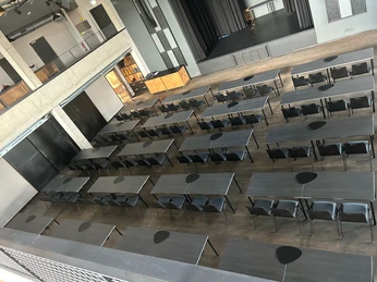 Parlamentarisch.jpg Großer Veranstaltungssaal mit grauen Tischen und Stühlen, Bühne vorne, Galerie im oberen Bereich.