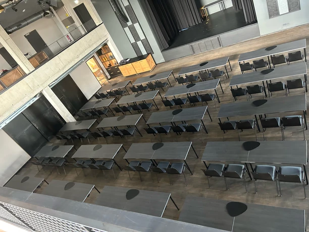 Parlamentarisch.jpg Großer Veranstaltungssaal mit grauen Tischen und Stühlen, Bühne vorne, Galerie im oberen Bereich.
