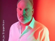 Mann mit Bart im weißen Hemd steht vor einem farbig beleuchteten Hintergrund.