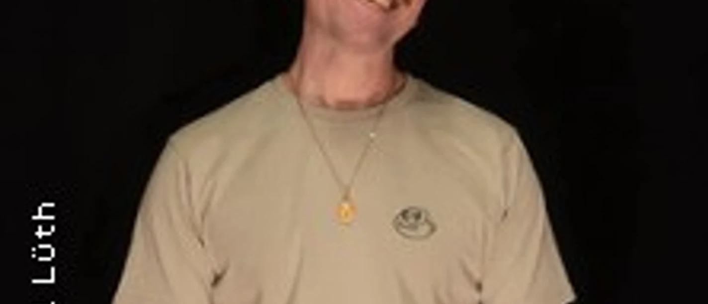 Eine Person mit Glatze und Bart, trägt ein beiges T-Shirt, lächelnd vor schwarzem Hintergrund.
