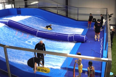 Surfanlage in Pinkys Surfhouse, Mettmann Surfschule mit blauer Indoor-Welle und mehreren Personen, die das Surfen üben.