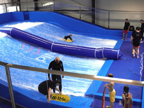 Surfanlage in Pinkys Surfhouse, Mettmann Surfschule mit blauer Indoor-Welle und mehreren Personen, die das Surfen üben.