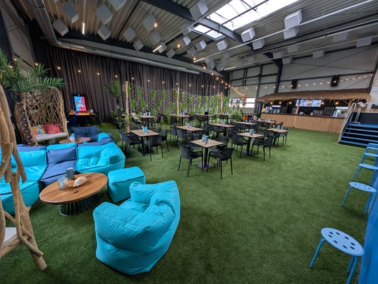 Lounge in Pinkys Surfhouse, Mettmann Großer Raum mit hellen Möbeln, bunten Sitzsäcken auf grünem Kunstrasen und einer Bar.