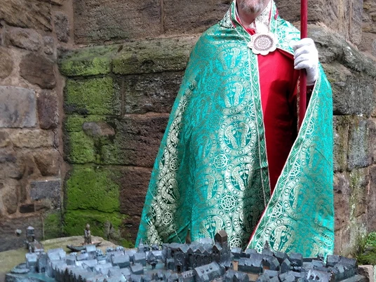 DVM _ Erlebnisfuehrung Mindener Dom (002).JPG Auf dem Bild sieht man Thomas Diekriede alias Bischof Wardenberg in einem grünen Mantel mit einer goldenen Mitra, einer spitz zulaufende Kopfbedeckung. Der Bischof steht am Dom hinter dem Stadtmodell aus Bronze. In der Hand hält er einen Hirtenstab.