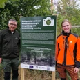 Fotospot Spiegelsberge Zwei Personen stehen im Grünen neben einem Schild zum Waldspendeprojekt Spiegelsberge.