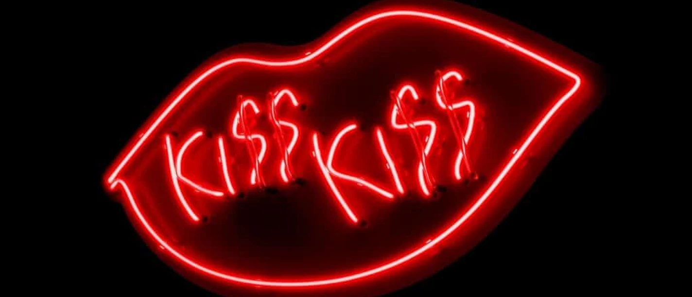 Leuchtende Neonlinien in Form von einem Mund mit "KISS KISS" in kräftigem Rot auf schwarzem Hintergrund.