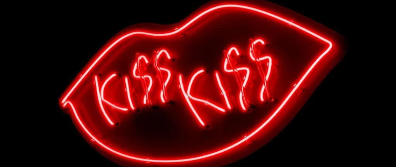 KISS KISS (c) Shu Lea Cheang.jpg Leuchtende Neonlinien in Form von einem Mund mit "KISS KISS" in kräftigem Rot auf schwarzem Hintergrund.