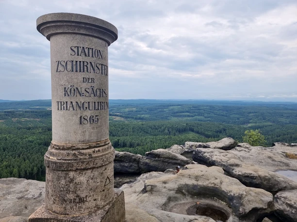 Stone column with inscription "Station Zschirnstein der Kön. Sächs. Triangulirung 1865" in front of a view of a forest landscape.