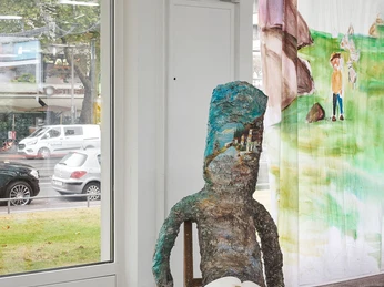 Amelie von Wulffen, Installationsansicht Eine surreale Skulptur sitzt auf einem Stuhl in einer Galerie, ein Skizzenbuch auf dem Schoß.A surreal sculpture sits on a chair in a gallery, a sketchbook on its lap.