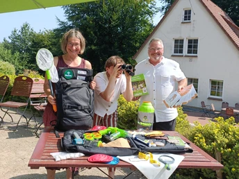 NFH Teutoburg NahTour Team c NaturFreunde NRW.jpg Drei Menschen stehen vor einem Tisch am Naturfreunde Haus Teutoburg und zeigen den Inhalt des Rucksacks