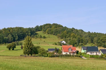 Landelijk tafereel met huizen, velden en beboste heuvels onder een blauwe lucht.