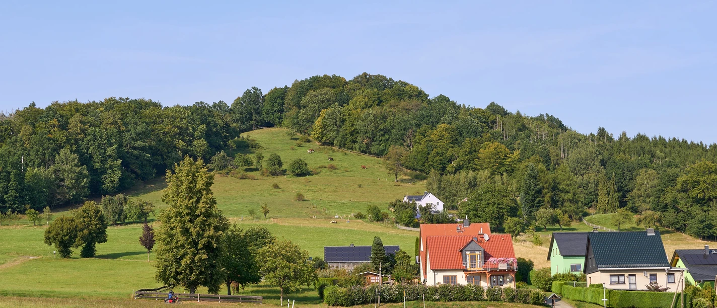 Landelijk tafereel met huizen, velden en beboste heuvels onder een blauwe lucht.