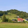 Gickelsberg Ländliche Szene mit Häusern, Feldern und bewaldeten Hügeln unter blauem Himmel.