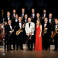 Eine Big Band in eleganter Abendkleidung posiert auf einer Bühne mit Blasinstrumenten.
