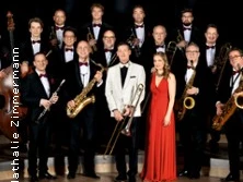 Eine Big Band in eleganter Abendkleidung posiert auf einer Bühne mit Blasinstrumenten.