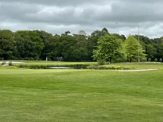 Rasenfläche des Oldenburgischen Golfclub Grüne Golfanlage mit Teich, zwei kleinen Brücken, Wasserfontäne und Bäumen unter grauem Himmel