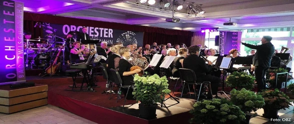 orchester-bad-zwischenahn Das Bild zeigt ein Orchester im Raum mit lila beleuchteter Bühne. Musiker spielen Blasinstrumente, während im Hintergrund Schlagzeuger zu sehen sind. Gäste sind an Tischen platziert. Der Raum ist geschmückt mit grünen Pflanzen, und der Schriftzug "Orchester Bad Zwischenahn" ist im Hintergrund sichtbar.