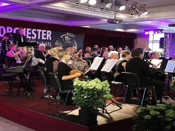 Das Bild zeigt ein Orchester im Raum mit lila beleuchteter Bühne. Musiker spielen Blasinstrumente, während im Hintergrund Schlagzeuger zu sehen sind. Gäste sind an Tischen platziert. Der Raum ist geschmückt mit grünen Pflanzen, und der Schriftzug "Orchester Bad Zwischenahn" ist im Hintergrund sichtbar.