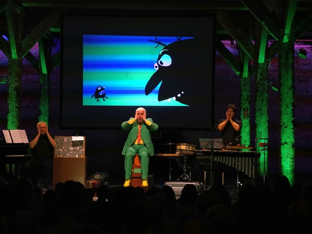 voices 2.jpg Bühnenauftritt mit drei Künstlern vor Leinwand, darauf bunte Cartoon-Figuren, Publikum zuschauend.