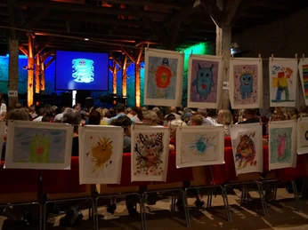 voices_kinderkonzert.jpg Kinderzeichnungen mit bunten Fantasiefiguren hängen vor Publikum in einem Konzertsaal.