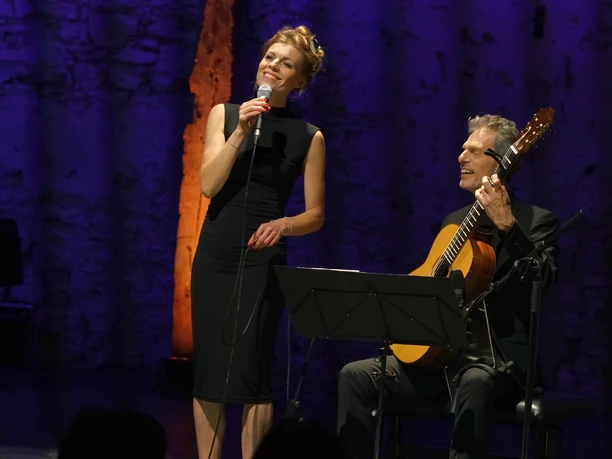 voices1.jpg Frau singt mit Mikrofon neben Gitarrist auf beleuchteter Bühne vor Publikum.