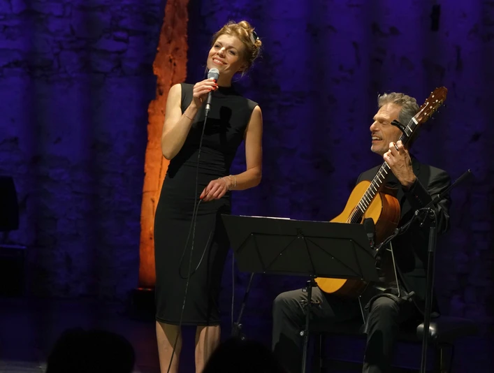 voices1.jpg Frau singt mit Mikrofon neben Gitarrist auf beleuchteter Bühne vor Publikum.