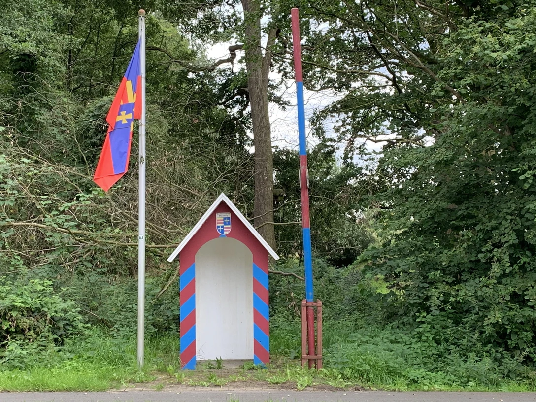 Grenzstation.jpg Kleine bunte Grenzstation mit rot-blauer Schranke und Flagge vor dicht bewachsenem Wald.