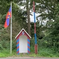 Grenzstation.jpg Kleine bunte Grenzstation mit rot-blauer Schranke und Flagge vor dicht bewachsenem Wald.