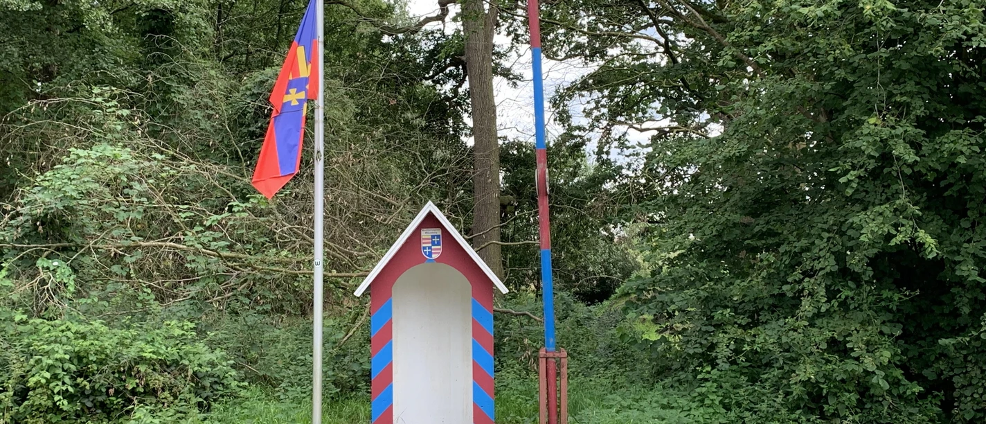 Grenzstation.jpg Kleine bunte Grenzstation mit rot-blauer Schranke und Flagge vor dicht bewachsenem Wald.