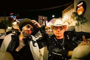 the-bosshoss-image.jpg Frontmen (Alec und Sascha) von The BossHoss sitzen mit Sonnenbrille und Cowboyhüten bei Nacht in einem Cabrio, im Hintergrund Häuser und Lichter