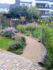 Gemeinschaftsgarten Ein kleiner, gepflasterter Weg führt durch einen farbenfrohen Garten mit Blumenbeeten und Sträuchern.
