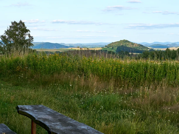 Wooden bench in the foreground with a view of green hills and a cloudy sky.