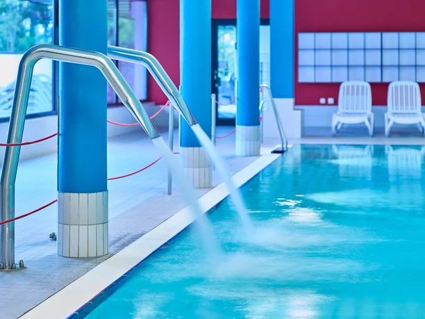 Nichtschwimmerbecken Oktopus Das Bild zeigt den Innenbereich eines Schwimmbads mit leuchtend blauen Säulen und einem ruhigen Wasserbecken. Im Hintergrund sind zwei weiße Liegestühle vor einer roten Wand sichtbar.