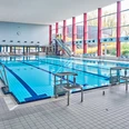 Schwimmerbecken Oktopus Ein großes, lichtdurchflutetes Schwimmbecken in einer modernen Halle. Üppige Fenster auf der rechten Seite lassen Tageslicht herein, während links Leitern den Einstieg ins Wasser ermöglichen. Im Hintergrund sind Wasserrutschen und Sitzgelegenheiten zu sehen, ideal für Sport und Entspannung.