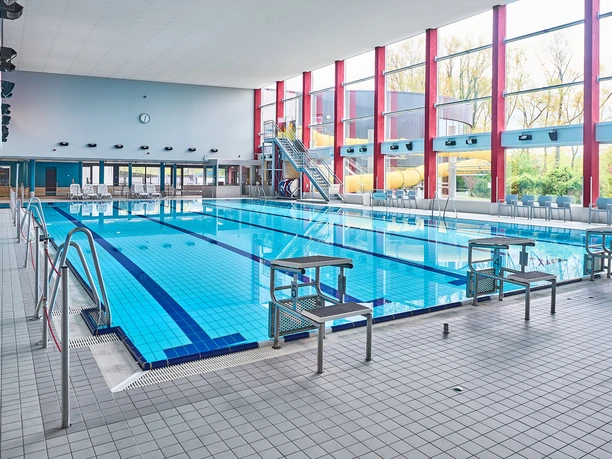 Schwimmerbecken Oktopus Ein großes, lichtdurchflutetes Schwimmbecken in einer modernen Halle. Üppige Fenster auf der rechten Seite lassen Tageslicht herein, während links Leitern den Einstieg ins Wasser ermöglichen. Im Hintergrund sind Wasserrutschen und Sitzgelegenheiten zu sehen, ideal für Sport und Entspannung.