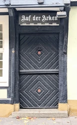 ©Bastian Ulrich - Haus auf der Arken 1 Schwarze Holztür mit kunstvollen Verzierungen unter einem weißem Schild mit der Aufschrift "Auf der Arken".