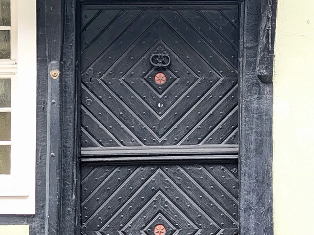©Bastian Ulrich - Haus auf der Arken 1 Schwarze Holztür mit kunstvollen Verzierungen unter einem weißem Schild mit der Aufschrift "Auf der Arken".
