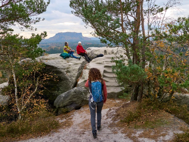 Vrouw loopt op pad naar twee mensen die op een rots zitten in een berglandschap.