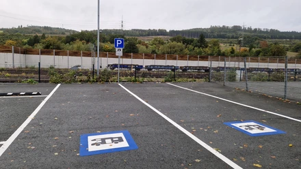 Ein Parkplatz mit Bodenmarkierungen für Wohnmobile, dahinter Gleise und eine vegetationsreiche Umgebung.