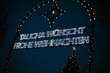 Leuchtendes Weihnachtsschild in Taucha bei Nacht, umrahmt von bunten Lichtern und einem Tannenbaum.