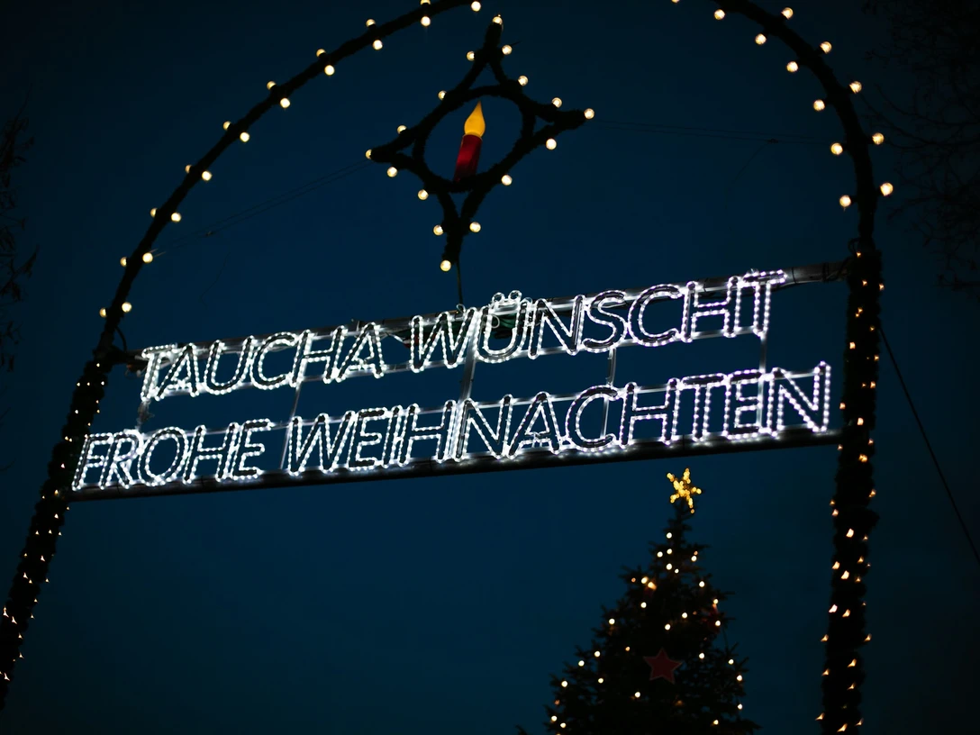 Weihnachtsmarkt Taucha - Weihnachten in der Region Leipzig Leuchtendes Weihnachtsschild in Taucha bei Nacht, umrahmt von bunten Lichtern und einem Tannenbaum.