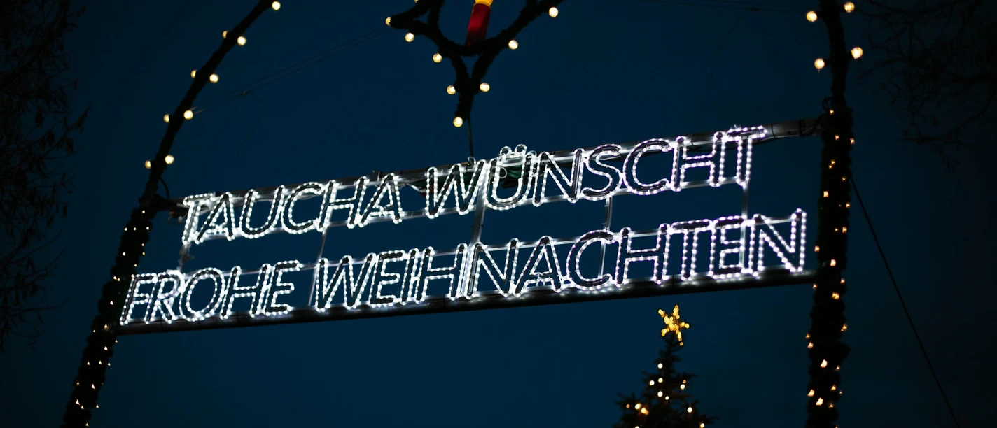 Weihnachtsmarkt Taucha - Weihnachten in der Region Leipzig Leuchtendes Weihnachtsschild in Taucha bei Nacht, umrahmt von bunten Lichtern und einem Tannenbaum.