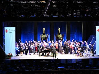 Rotkreuz-Benefiz-Matinee der Oper Köln Ein Orchester und Chor präsentieren ein Konzert auf großer Bühne, umgeben von skulpturalen Pferdeplastiken.An orchestra and choir present a concert on a large stage, surrounded by sculptures of horses.