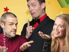 Drei Personen in festlicher Kleidung mit humorvollen Gesten vor buntem Hintergrund.