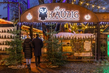 Wintermarkt bei Herr Kauzig - Weihnachten in Leipzig Zwei Personen betreten den Eingang zum kauzigen Wintermarkt im Biergarten von Herrn Kauzig, links und rechts vom Eingang stehen zwei Tannenbäume und im Hintergrund sind festlich beleuchtete Stände zu sehen