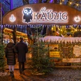 Wintermarkt bei Herr Kauzig - Weihnachten in Leipzig Zwei Personen betreten den Eingang zum kauzigen Wintermarkt im Biergarten von Herrn Kauzig, links und rechts vom Eingang stehen zwei Tannenbäume und im Hintergrund sind festlich beleuchtete Stände zu sehen