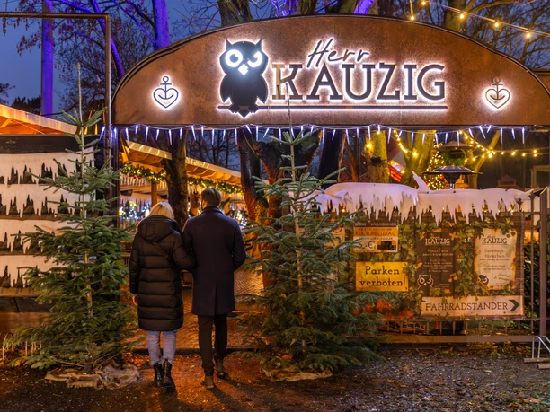 Wintermarkt bei Herr Kauzig - Weihnachten in Leipzig Zwei Personen betreten den Eingang zum kauzigen Wintermarkt im Biergarten von Herrn Kauzig, links und rechts vom Eingang stehen zwei Tannenbäume und im Hintergrund sind festlich beleuchtete Stände zu sehen