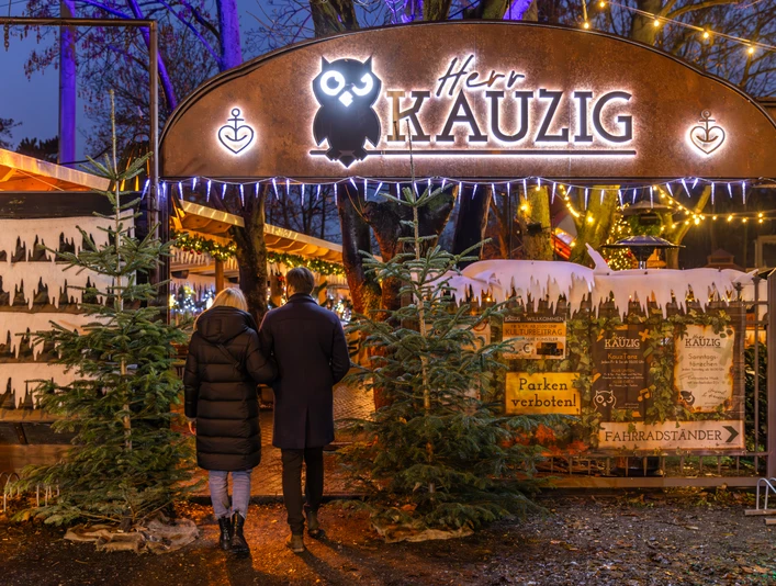 Wintermarkt bei Herr Kauzig - Weihnachten in Leipzig Zwei Personen betreten den Eingang zum kauzigen Wintermarkt im Biergarten von Herrn Kauzig, links und rechts vom Eingang stehen zwei Tannenbäume und im Hintergrund sind festlich beleuchtete Stände zu sehen