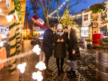 Wintermarkt bei Herr Kauzig - Weihnachten in Leipzig Drei Personen stehen auf dem Gelände des kauzigen Wintermarktes während im Hintergrund die festlich beleuchteten Verkaufsstände und der große Weihnachtsbaum zu sehen sind